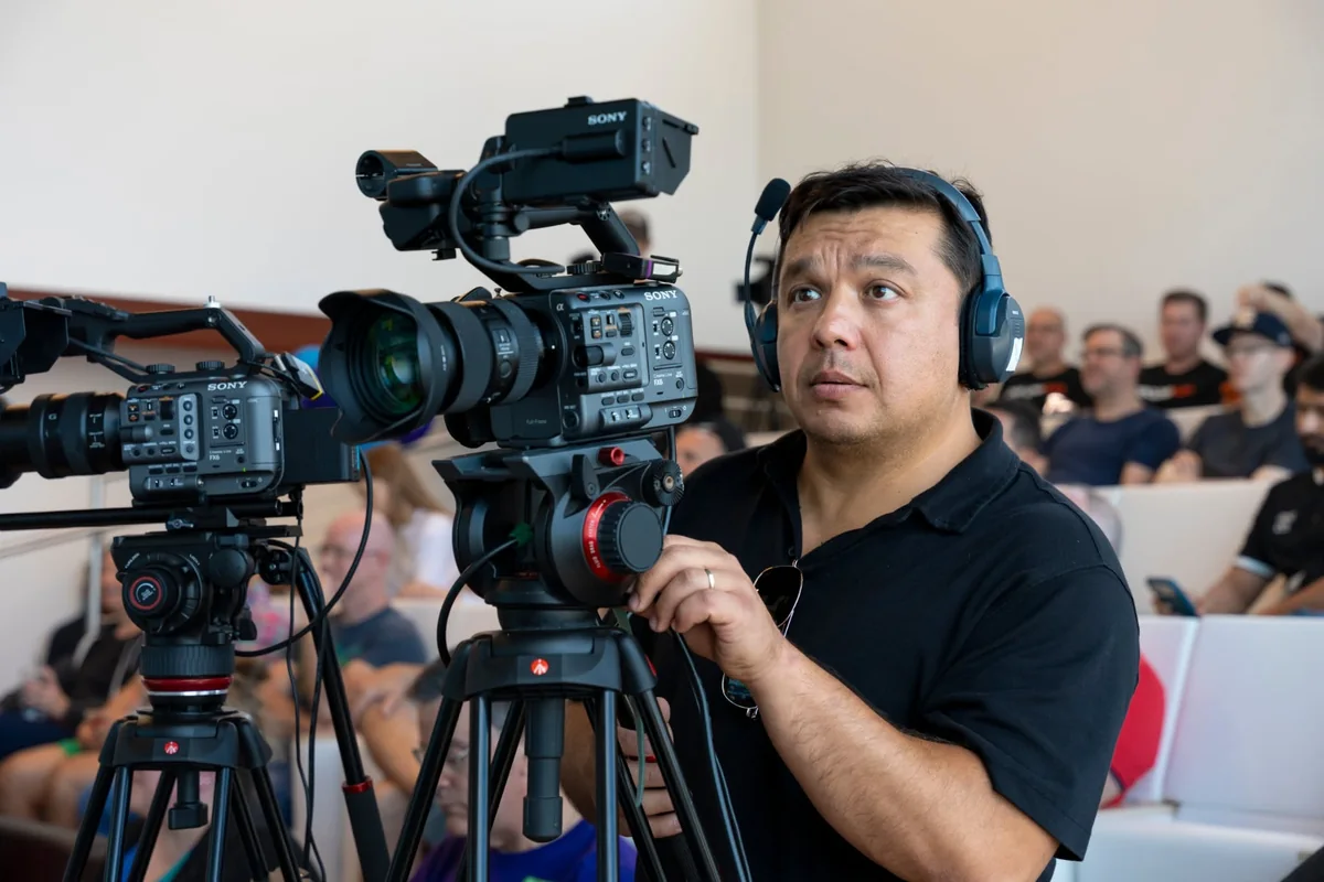 Camera operator wearing headphones beside two cameras, with a seated audience behind him.