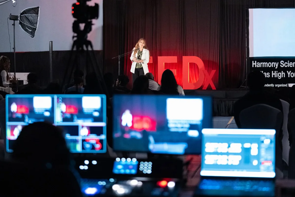 Production monitors showing camera feeds, with a speaker on the Harmony TEDx stage beyond them.