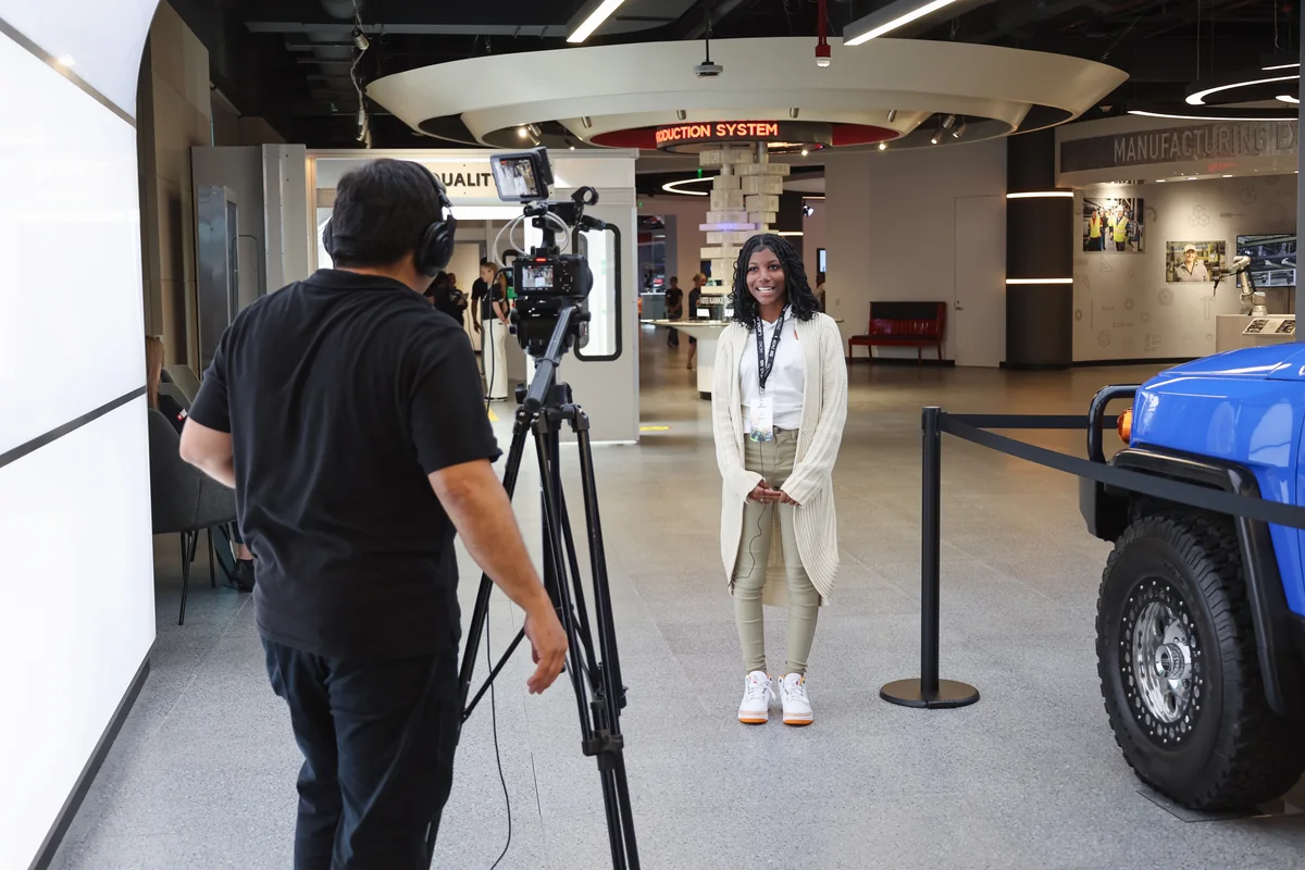 A camera operator records an interview with a participant beside a vehicle display during the Lexus visit.