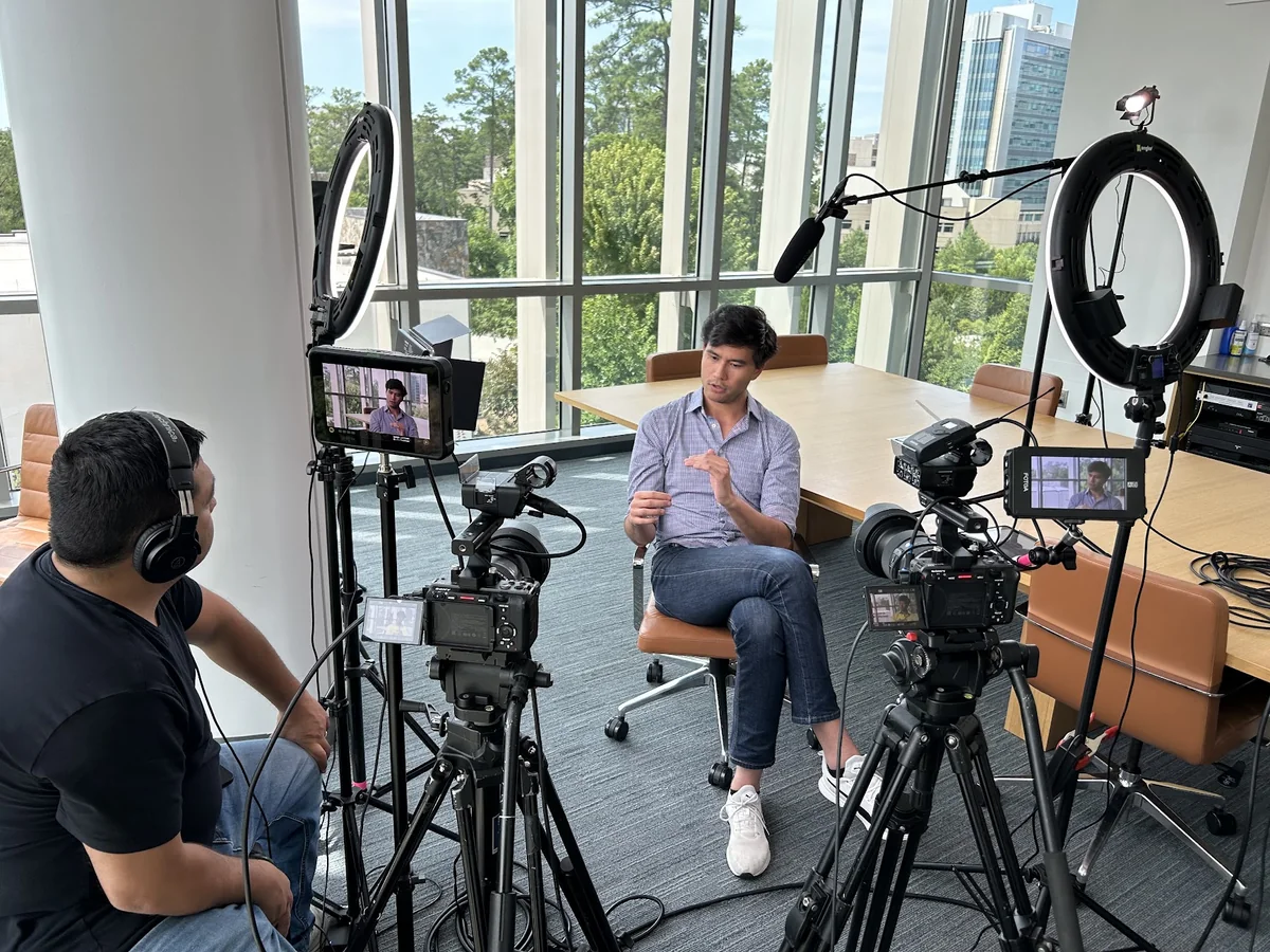A crew member listens during a seated interview at Duke, with two cameras, lights, and a boom microphone around the interviewee.