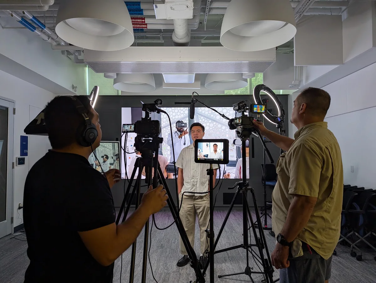 Two crew members operate cameras around an interviewee at Duke, with virtual reality imagery on the display behind him.